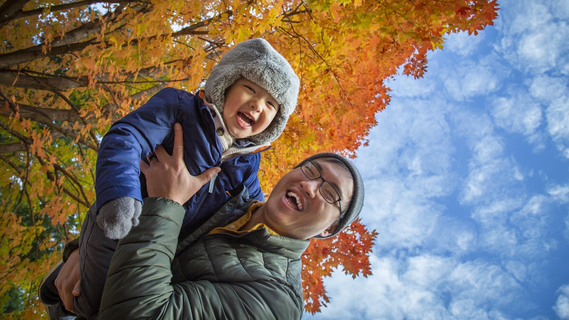All of Us Hmong father holding young son outdoors, both smiling at camera in front of maple tree with orange leaves